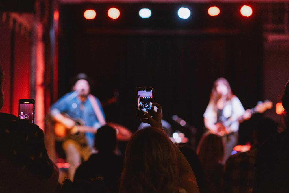 Fans on the floor at the Music Hall with hands raised enjoying a performance on stage.