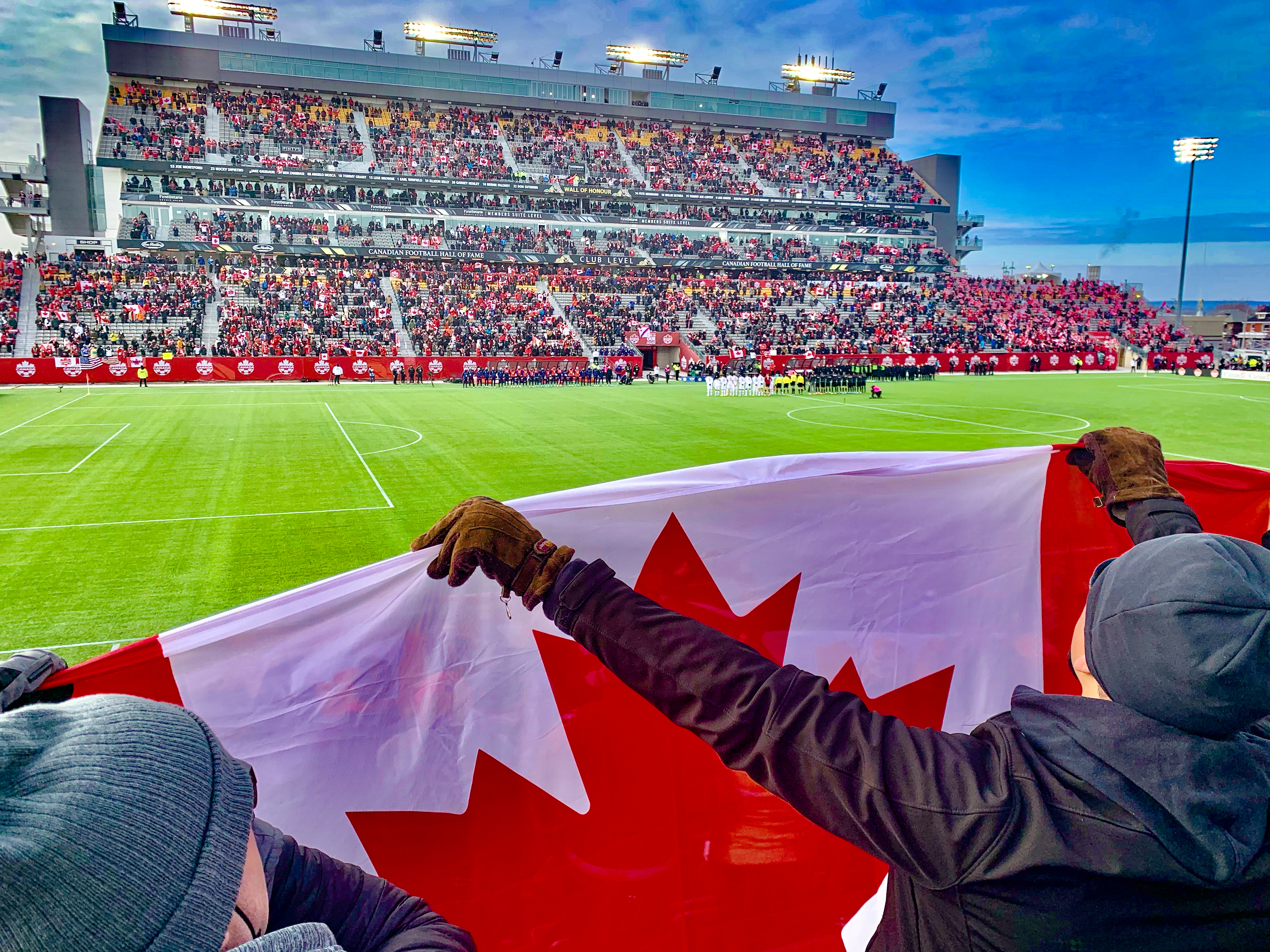 Hamilton Stadium from the perspective of the audience. People are wearing red and white, with the main subject of the photo being the sports field and an individual holding up the Canadian flag.