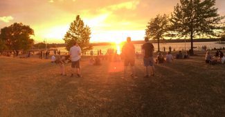 People sitting and standing on grass in front of sun setting over Hamilton Harbour