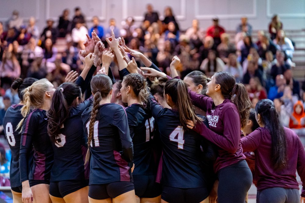 McMaster Women's Volleyball team huddle on court in front of full crowd in gymnasium.