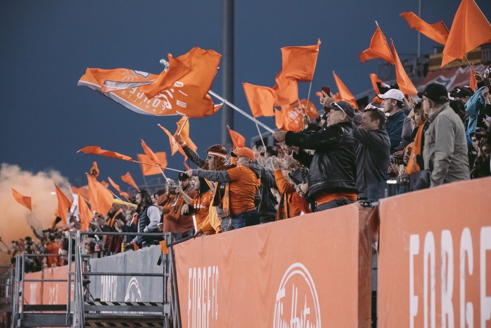 Forge FC fans in the stands cheering and waving flag.