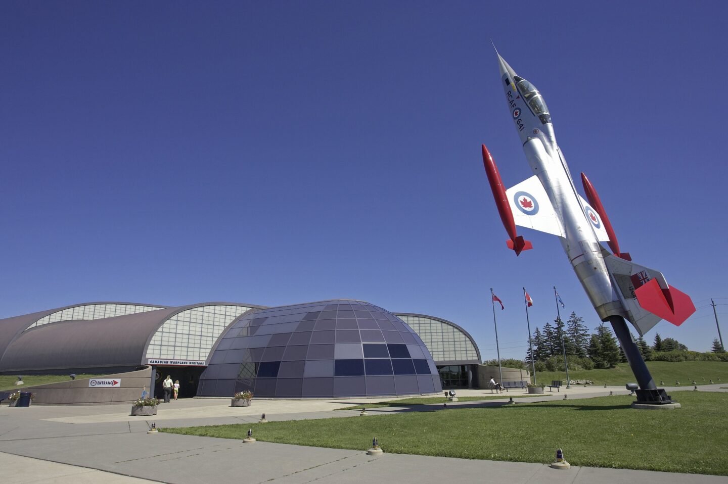 A fighter jet displayed upright outside the Canadian Warplane Heritage Museum in winter, surrounded by snow-covered shrubs with flags flying above the curved hangar building under a clear blue sky.