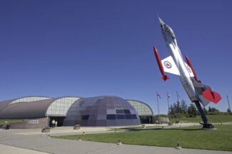 A fighter jet displayed upright outside the Canadian Warplane Heritage Museum in winter, surrounded by snow-covered shrubs with flags flying above the curved hangar building under a clear blue sky.