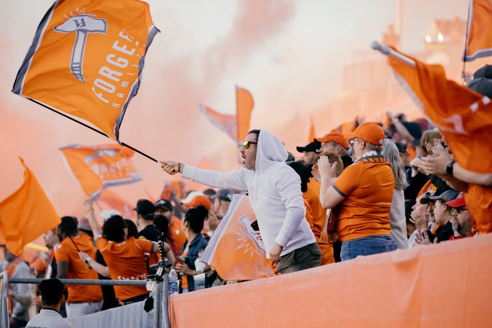 Full stand of Forge FC fans in orange waving the team flag.