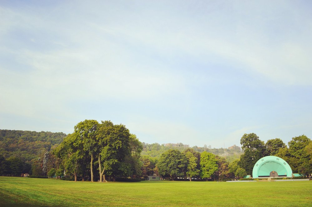 Aqua green bandshell stage set in the distance of a park. Surrounded by green grass and trees.