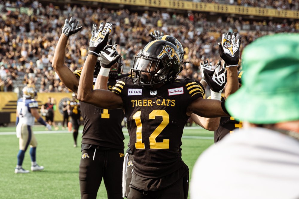 Ticats players with arms raised on the field looking at the camera.