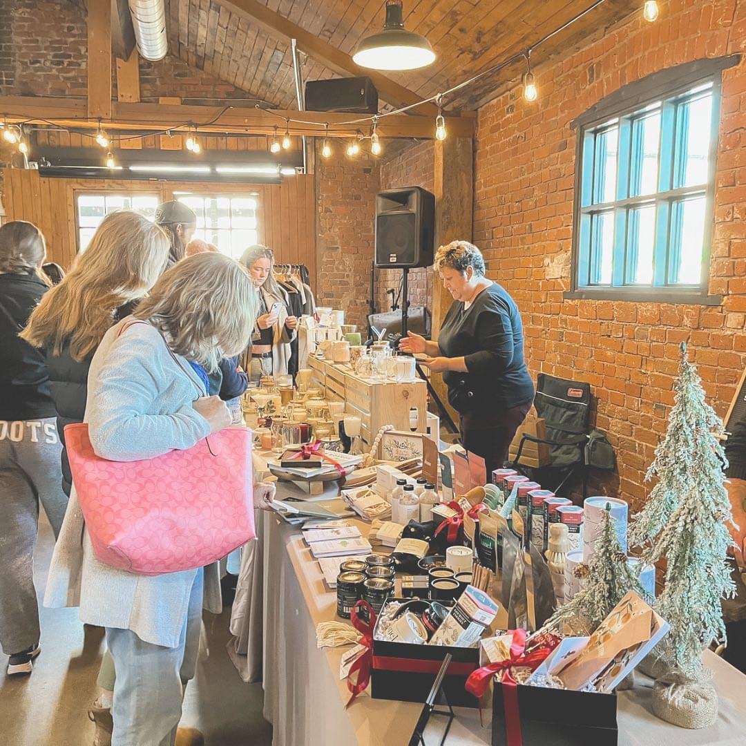 People browsing a holiday market inside a rustic brick building with string lights, where vendors display handmade gifts, candles, and local products on decorated tables.