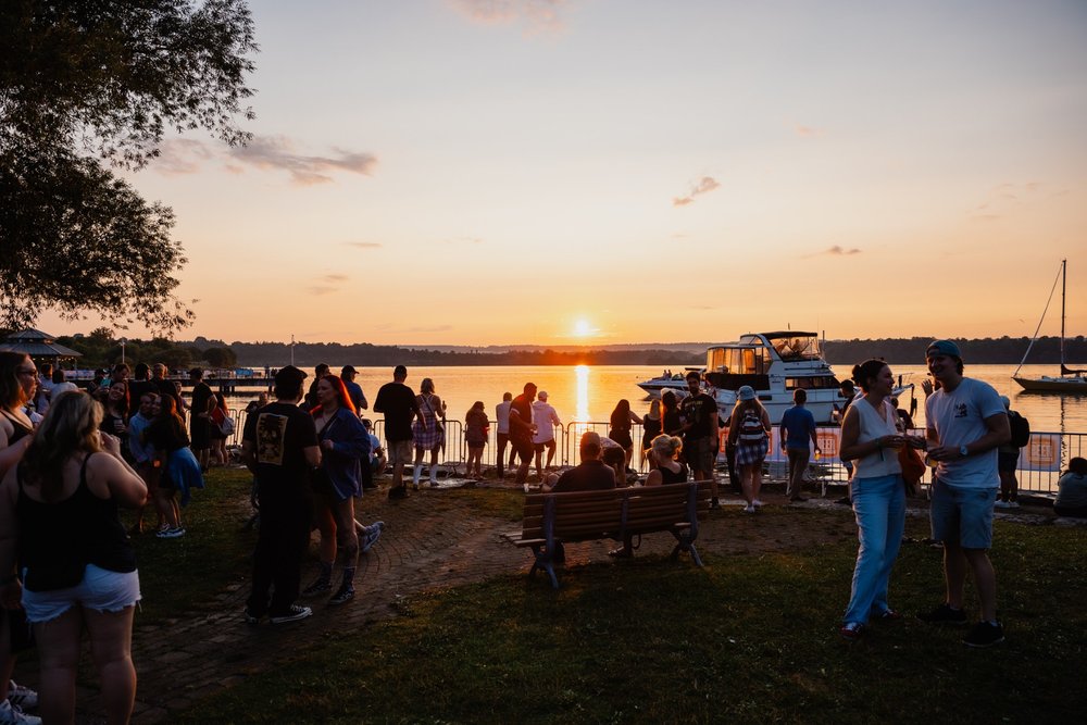 People sitting and standing on grass in front of sun setting over Hamilton Harbour