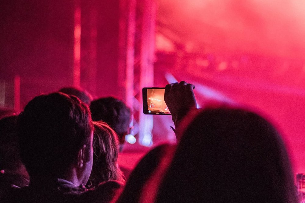 Individual in crowd taking photo of band playing on stage at concert.