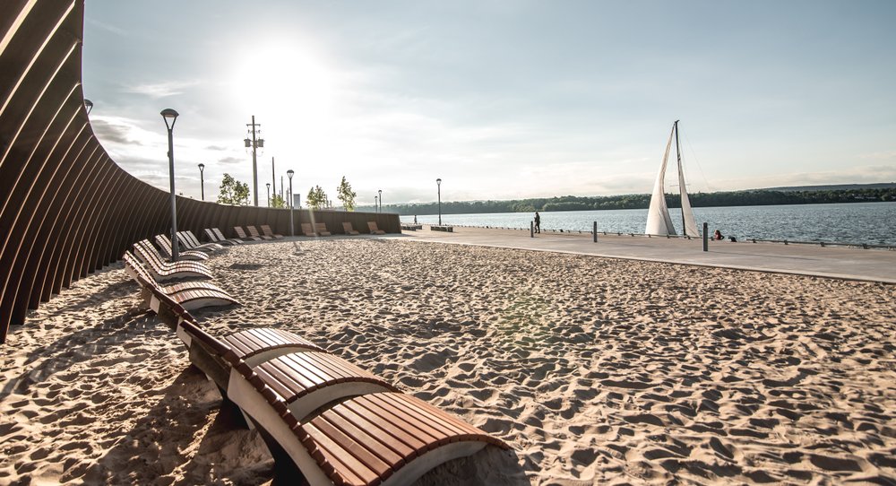 Sandy beach with wooden lounge chairs face the glistening waterfront. A white sail boats floats by.