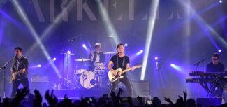 Canadian rock band Arkells performing on stage under bright spotlights, with the lead singer playing guitar at centre stage, drummer and keyboardist behind him, and a cheering crowd in the foreground.