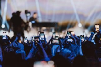Concert crowd holding up smartphones to record a live performance under bright blue and white stage lights, with the performer blurred in the background. The scene captures the energy and excitement of a large audience at a modern music event.