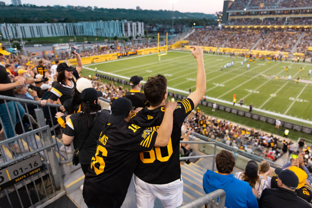 Fans looking onto the field at Hamilton Stadium during a Ticats game and cheering.