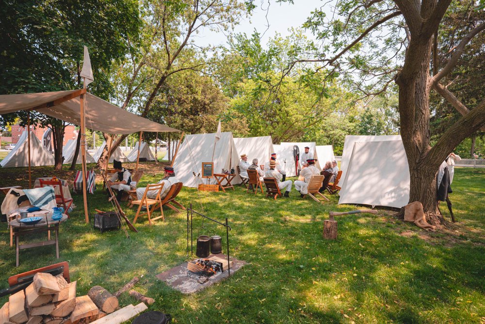 Costumed historical re-enactors in period military dress gathered around a campfire at a living history encampment, with white canvas tents set up across a grassy field under mature trees.