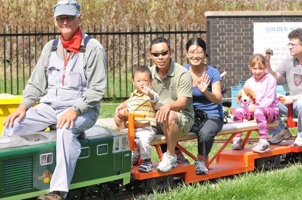 Conductor riding a green miniature train with kids and adult passengers enjoying the ride.
