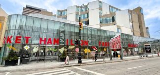 Exterior view of the Hamilton Farmers’ Market in downtown Hamilton, showing its modern glass façade with large red lettering spelling “HAMILTON FARMERS MARKET.” Pedestrians walk by on the sidewalk, and colorful produce illustrations decorate the windows beneath a cloudy sky.