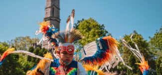 A dancer in traditional Indigenous regalia performs at an outdoor event. The dancer wears a vibrant outfit adorned with colorful feathers in shades of blue, orange, and white, along with intricate beadwork and ribbons. A large feathered headdress and arm accessories accentuate the movement. A historic stone monument and lush green trees are visible in the background under a bright blue sky.