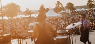 A musician performs on stage at an outdoor festival during golden hour, with a large crowd gathered on the grass under trees and white tents. The warm sunlight creates a glowing, energetic atmosphere as people enjoy live music near a waterfront.