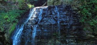 A natural waterfall cascades down layered rock formations surrounded by lush green vegetation and forest. The water glistens as it flows gently over the dark stone, creating a tranquil, shaded scene in the woods.